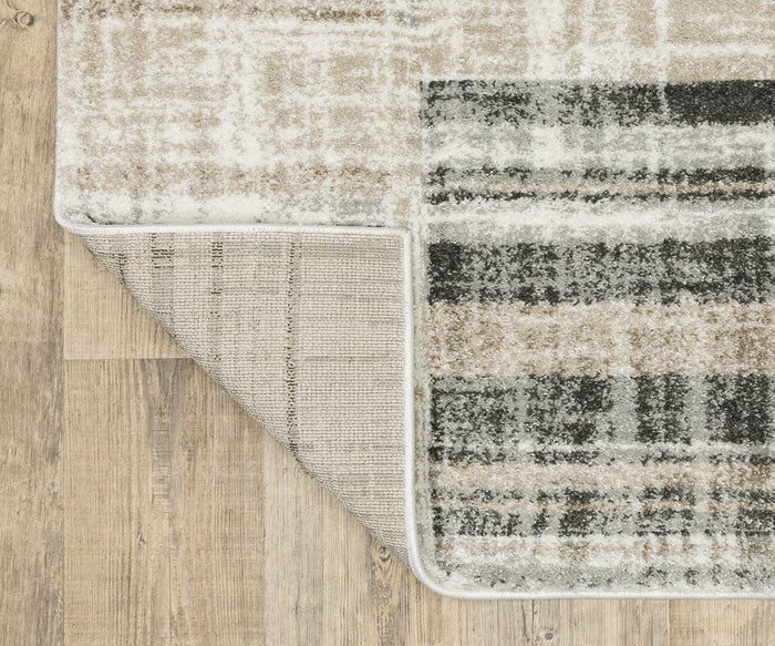A corner of the Oriental Weavers Cambria / 55X Rug, featuring a beige, black, and gray plaid pattern, is flipped over to reveal its durable woven underside made from polypropylene yarn. The high-pile rug rests on a light brown wooden floor.