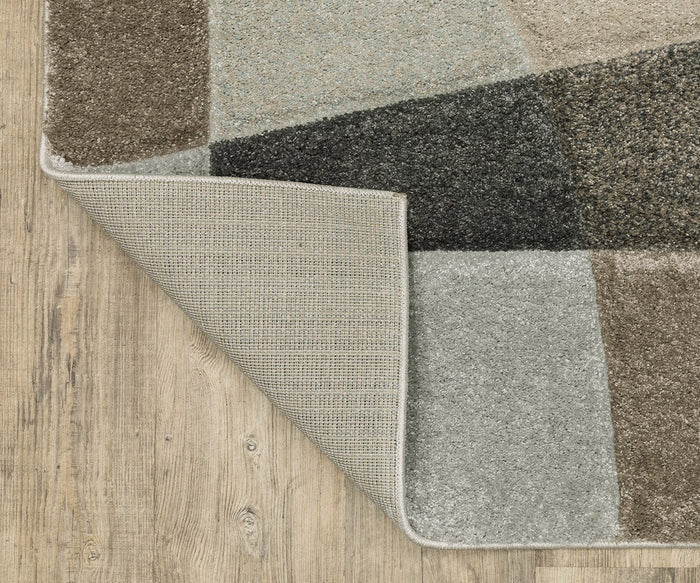 A corner of the Oriental Weavers Cambria / 84J Rug is flipped over to reveal its non-slip backing. The high-pile rug rests on a light wood floor, showcasing intricate geometric motifs.