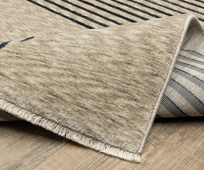 A corner of the Oriental Weavers Capella / CAP04 Rug is turned up, revealing the underside. This beige and black striped space-dyed polyester rug from the Capella Collection lies on a wooden floor, with a short fringe along the edge that accentuates its modern geometric designs.
