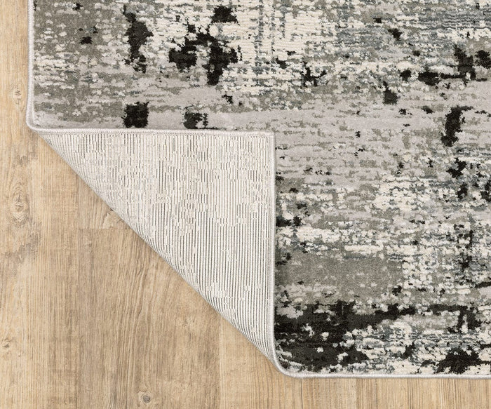The Oriental Weavers Caravan 4153Q rug features a rectangular shape with a muted gray and black abstract, nature-inspired pattern, pictured here atop a light wood floor. Part of the Caravan Collection, it is crafted from space-dyed polyester for subtle color variation. One corner is turned up to reveal its light gray underside.
