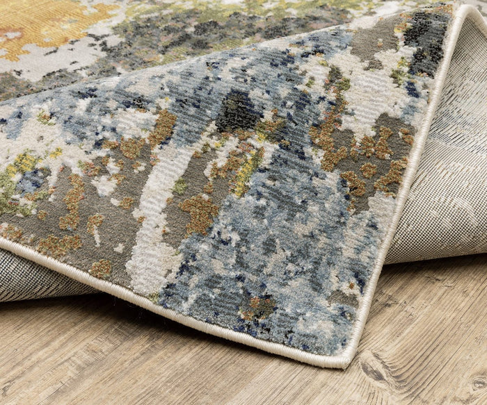 A close-up of the Oriental Weavers Caravan / 502X Rug showcases its abstract blue, gray, gold, and cream nature-inspired design. Expertly crafted from space-dyed polyester for an elegant look, this rug is partially folded on a light wooden floor.
