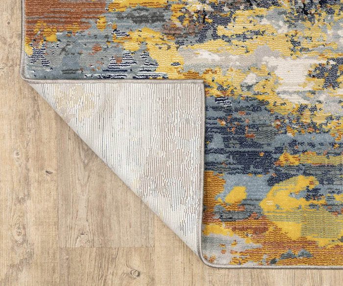 The Oriental Weavers Caravan / 530V Rug, crafted from space-dyed polyester and adorned with a nature-inspired abstract pattern in yellow, blue, gray, and brown accents, is pictured partly turned up at one corner to reveal its light, textured underside on a wooden floor. This vibrant rug from the Caravan Collection offers both enduring durability and eye-catching style for any room.