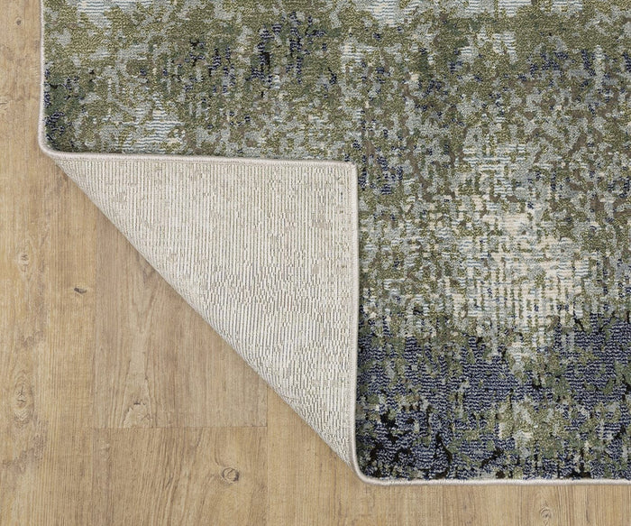 A corner of the Oriental Weavers Caravan 5571H Rug is folded over on a light wood floor, revealing the contrasting woven backing and the rug’s blue, green, and gray abstract nature-inspired pattern crafted from space-dyed polyester.