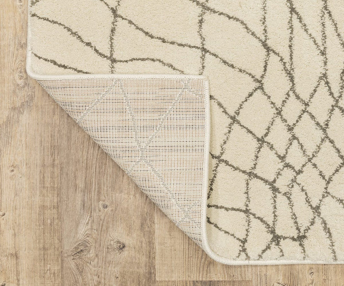 The Oriental Weavers Carson / 738B Rug, featuring a contemporary cream color with a gray abstract line pattern, is displayed on a light wood floor. One corner of the rug is flipped over, revealing its textured backing and emphasizing its durable polypropylene construction.