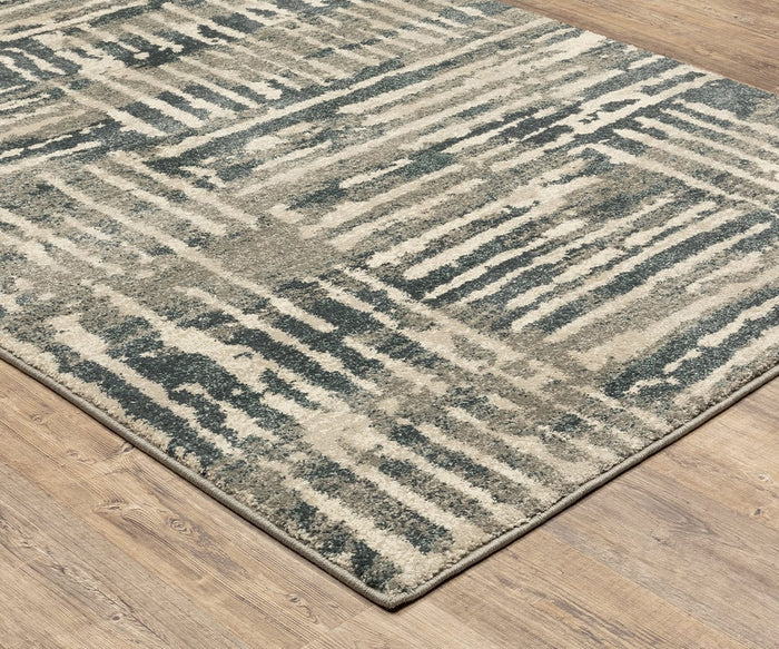 The Oriental Weavers Carson / 745C Rug is a contemporary rectangular area rug from the Carson Collection, showcasing an abstract striped design in tones of beige, gray, and blue. Placed on a light wood floor, its plush texture provides both comfort and modern style to any room.