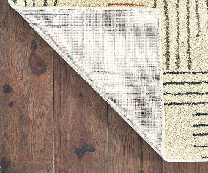 The Oriental Weavers Carson / 9663A Rug, featuring a beige pattern, is shown with one corner flipped over to reveal the backing. This contemporary rug rests on a brown wooden floor with visible grain and knots, bringing an inviting touch of plush comfort to the space.