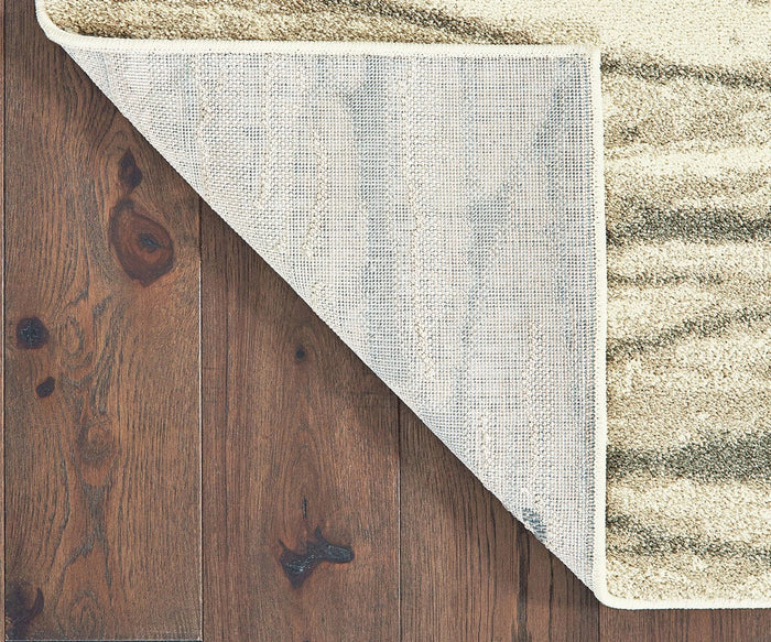 The Oriental Weavers Carson / 9671C Rug, featuring a beige patterned contemporary design with one corner flipped to show its non-slip backing, rests on a dark brown wooden floor with prominent grain and knots. Crafted from durable polypropylene, this rug offers a perfect combination of style and resilience for everyday living.