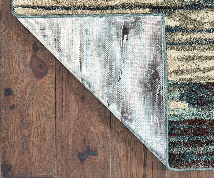 A corner of the Oriental Weavers Carson / 9675A Rug is flipped over to reveal the underside, resting on a wooden floor with visible grain and knots.