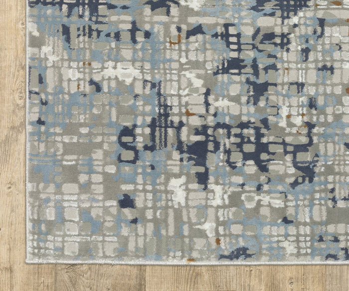 The Oriental Weavers Easton / 11E Rug, featuring gray, blue, white, and subtle brown accents, is displayed on a light wood floor. This durable power-loomed area rug showcases irregular, overlapping grid-like shapes and varied shading for a striking abstract pattern.