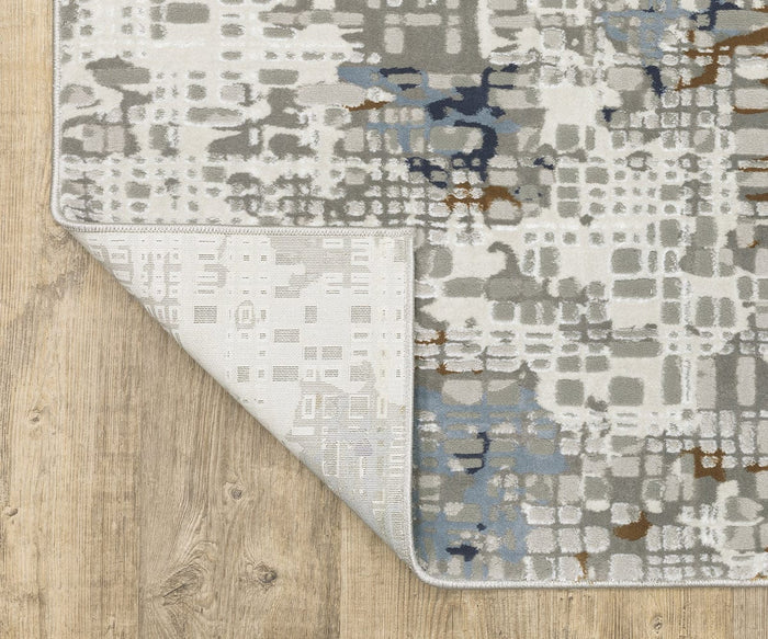 The Oriental Weavers Easton / 11E Rug is a modern abstract area rug featuring gray, blue, and brown patterns on a light background, displayed here on a wooden floor. This durable, power-loomed rug showcases artistic abstract designs throughout, with one corner flipped to reveal the underside.