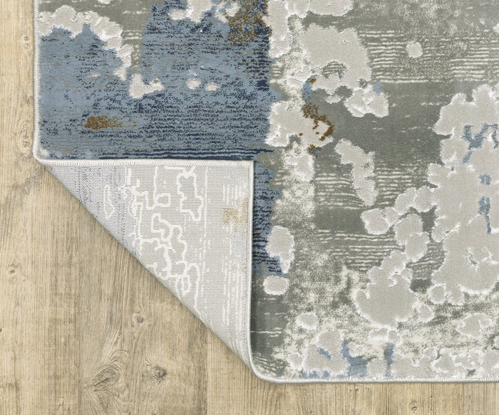 The Oriental Weavers Easton 4518X Rug from the Easton Collection, featuring modern abstract patterns in blue, gray, and white, is displayed on a light wood floor. One corner of this durable power-loomed rug is flipped over to reveal its underside and neatly stitched edges.