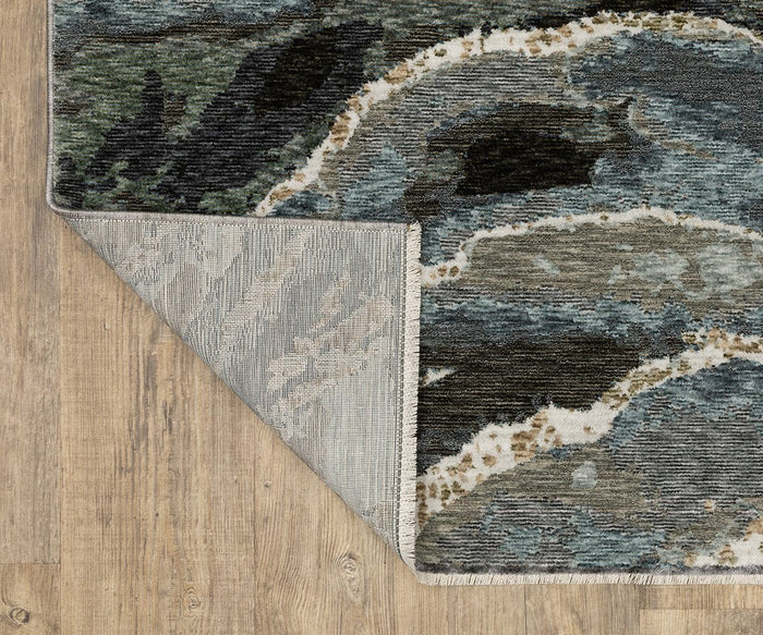 A corner of an Oriental Weavers Echo ECH12 Rug, power-loomed and made in Egypt, is flipped over to reveal its underside. The blue and gray patterned rug rests on a light brown wooden floor.