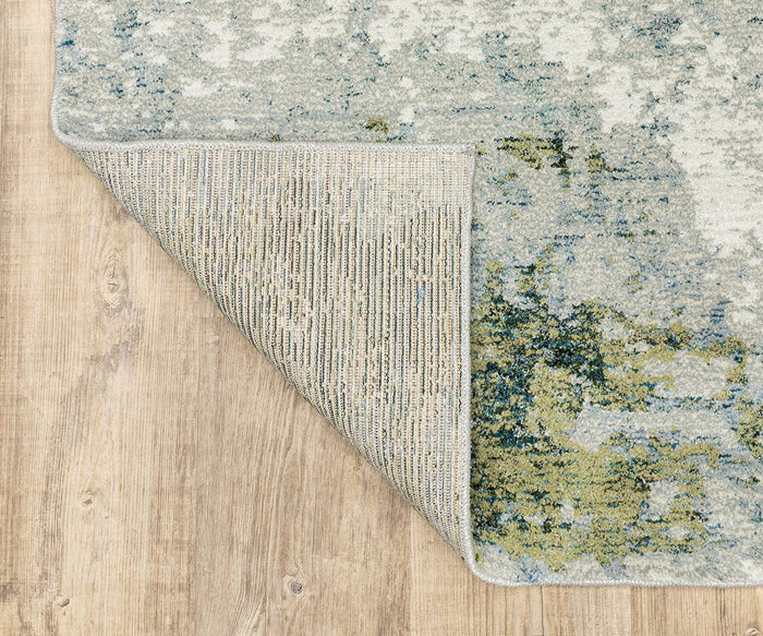 A corner of the Oriental Weavers Evolution / 8039E Rug is flipped over on a light wood floor, revealing its textured underside and an abstract blue, green, and beige design on top. Crafted from durable nylon, this easy-to-maintain rug seamlessly combines style with practicality.