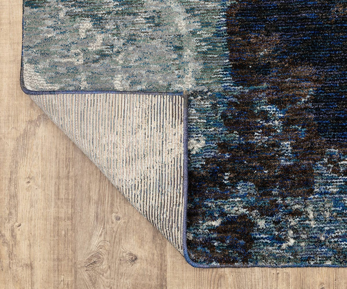 The Oriental Weavers Evolution / 962A Rug, featuring a blue and gray pattern, is shown with one corner flipped over to display the underside as it rests on a light wooden floor. Crafted from durable nylon, this rug combines style and practicality for everyday living and is easy to maintain.