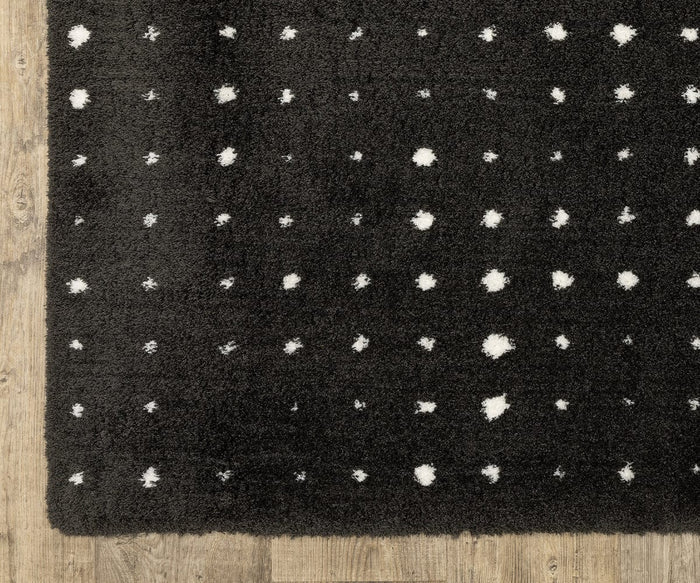 The photo features the Oriental Weavers Galileo / GAL04 Rug—a rectangular black plush rug adorned with scattered small white dots—positioned on a wooden floor. The image highlights the corner of the rug, emphasizing its soft texture and modern, minimalist design.