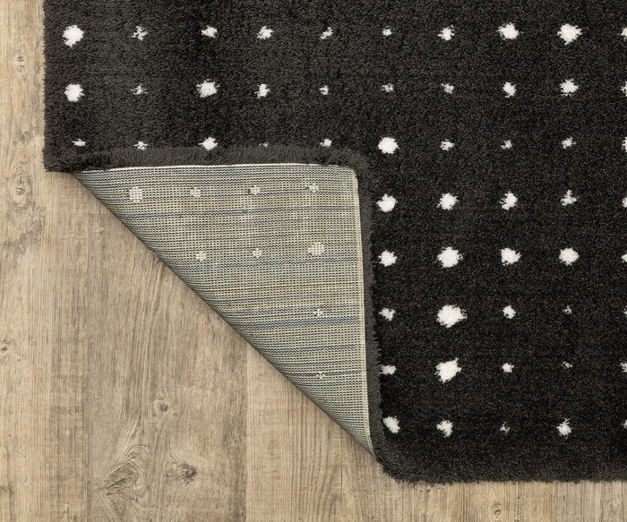 An Oriental Weavers Galileo / GAL04 Rug, featuring a contemporary black design with scattered white dots, is placed on a wooden floor. One corner of the plush rug is flipped over, revealing its textured gray underside.