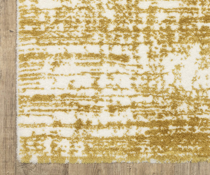 A close-up of the Oriental Weavers Galileo / GAL10 Rug showcases its soft, textured appearance and plush, dense pile. The rug features a contemporary gold and white abstract pattern on a wooden floor, with a slightly uneven edge adding to its modern appeal.