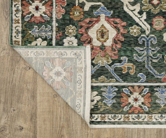 An Oriental Weavers Hastings / HA02B Rug featuring decorative floral patterns in green, red, blue, and cream is displayed on a wooden floor; the corner of this multicolor area rug is folded over to show its underside and stitched edges.