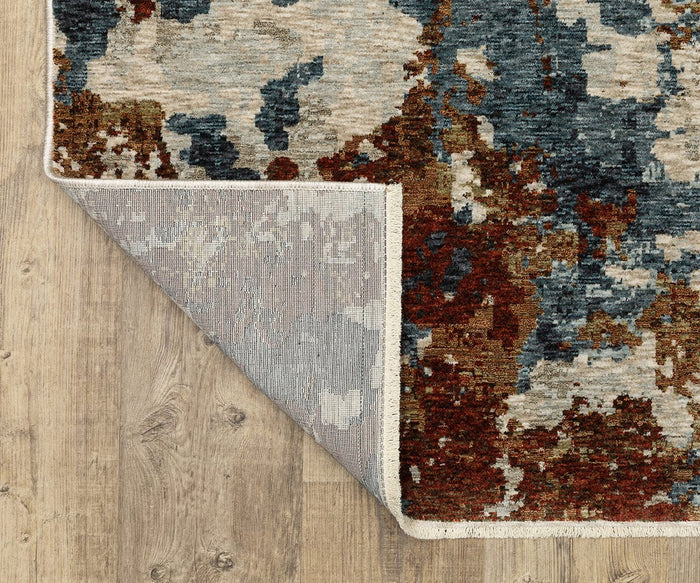 A close-up view of the rectangular Oriental Weavers Hayden / HAY01 Rug with its corner flipped to reveal the backing. This space-dyed polyester rug rests on a wooden floor and showcases modern abstract patterns in blue, rust, gray, and beige tones.