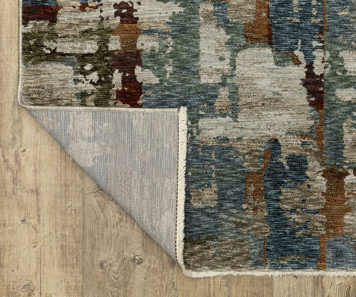 A corner of the Oriental Weavers Hayden / HAY02 Rug is folded over, revealing the underside, on a light wood floor. This rug features various shades of blue, green, brown, and beige in a textured space-dyed polyester design.