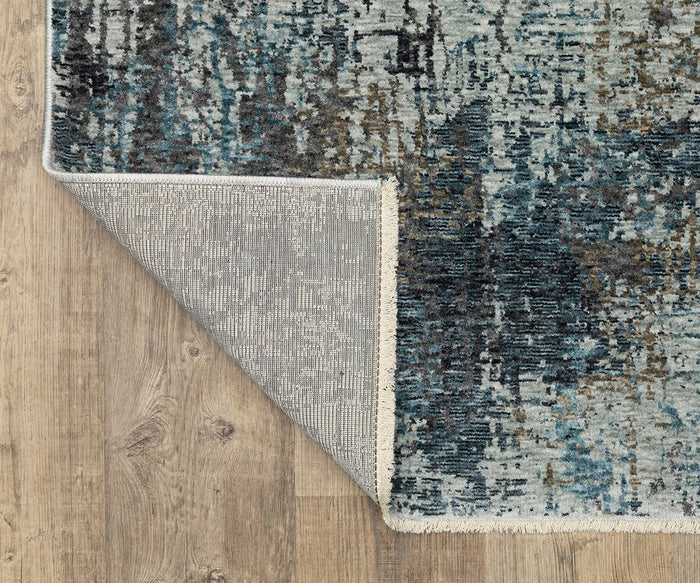 A corner of the Oriental Weavers Hayden HAY03 Rug, featuring its blue and gray abstract pattern, is flipped over to display the textured underside as it rests on a light wood floor.