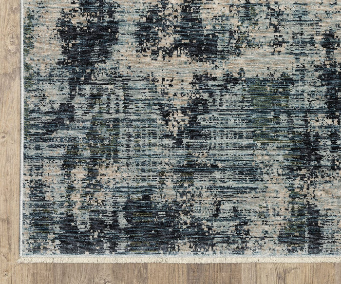 A close-up of the Oriental Weavers Hayden HAY04 Rug, a space-dyed polyester piece showcasing an abstract pattern in blue, beige, and gray tones on a wooden floor. This textured rug is accented with a light fringe along the edge.