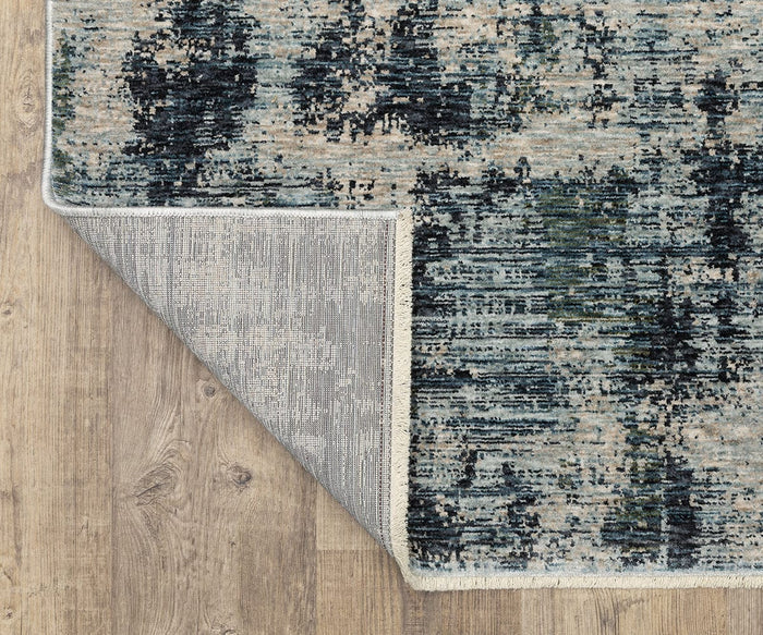 A close-up of the Oriental Weavers Hayden HAY04 Rug, featuring a blue and beige abstract pattern on a wooden floor, with one corner turned over to reveal the textured underside and showcase the space-dyed polyester’s intricate design.