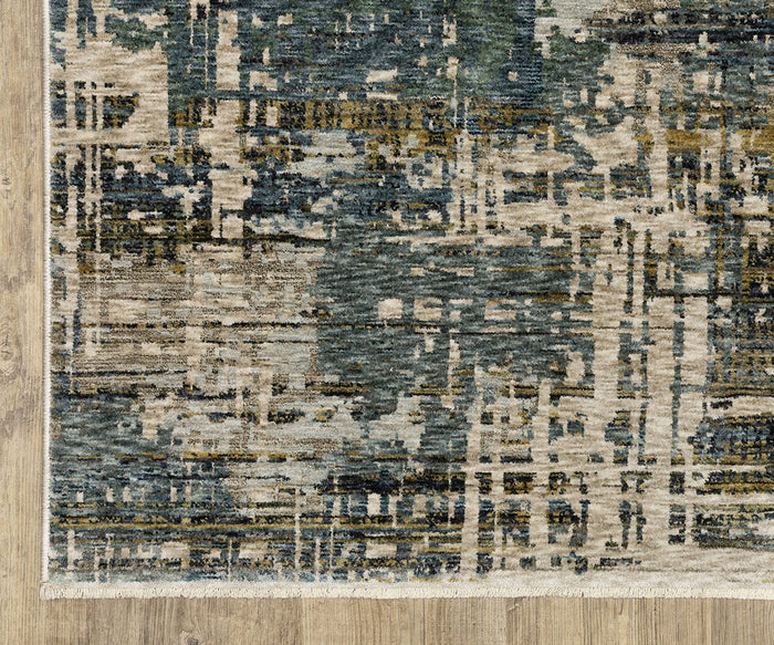 A close-up of the Oriental Weavers Hayden HAY05 Rug, a modern space-dyed polyester rug featuring an abstract pattern in blue, beige, and gold tones, placed on a light wood floor. The rug showcases a textured appearance with visible fringe along the edge.