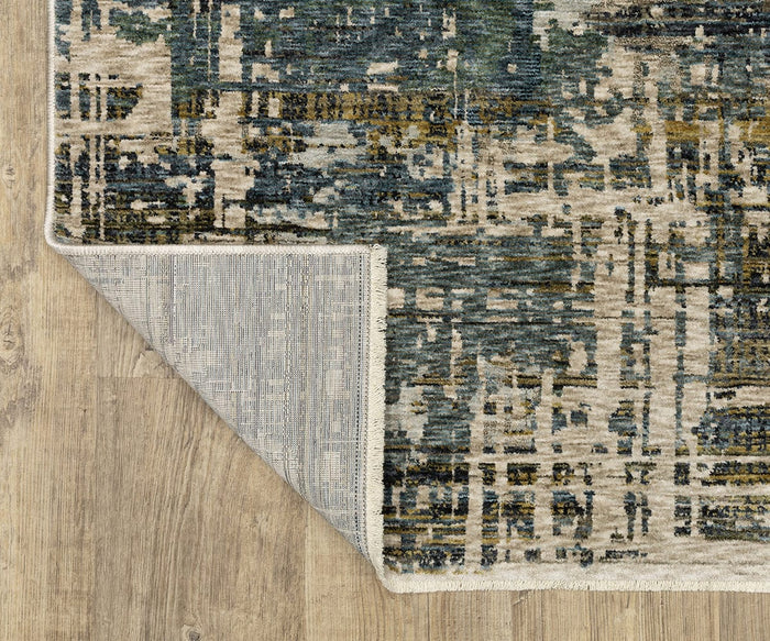 A modern space-dyed polyester rug, the Oriental Weavers Hayden HAY05, featuring abstract blue, beige, and olive patterns, lies on a wooden floor. One corner is flipped over, revealing the Hayden HAY05’s textured underside.