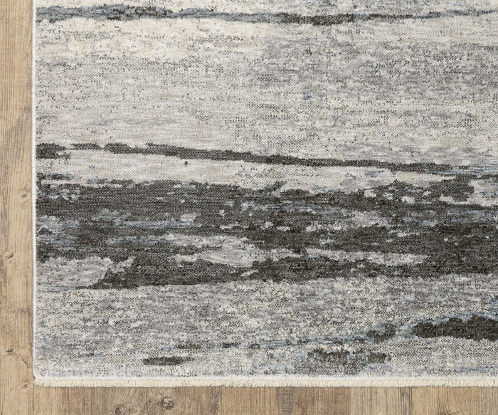 An Oriental Weavers Hayden / HAY07 Rug, featuring an abstract gray and black brushstroke pattern, is displayed on a wooden floor. Made from space-dyed polyester, the rug’s edge reveals its woven texture and short fringe.