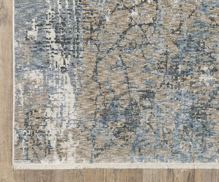 A close-up of the Oriental Weavers Hayden / HAY08 Rug, featuring an abstract pattern in blue, beige, and gray tones on a wooden floor. Crafted from space-dyed polyester, this area rug is finished with visible fringe on one edge for added texture.