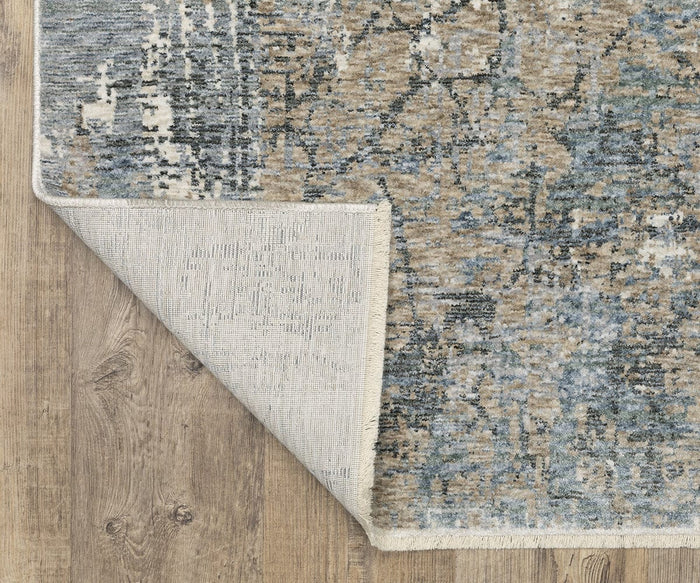 A corner of the Oriental Weavers Hayden / HAY08 Rug, crafted from space-dyed polyester, is folded over to reveal its backing. This abstract design features blue, beige, and gray patterns and is displayed on a wooden floor.