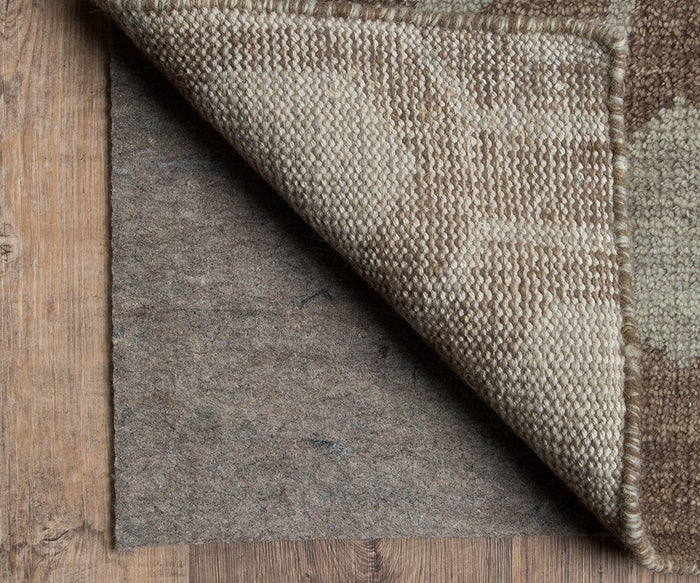 A corner of a patterned rug is lifted to reveal the Oriental Weavers Luxehold / 0005E Rug Pad underneath, both resting on a wooden floor.