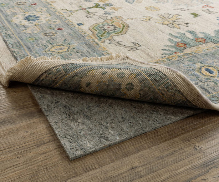 A corner of a patterned rug is lifted to reveal the Oriental Weavers Luxehold / 0005E Rug Pad underneath on a wooden floor. The rug features blue, beige, and green designs, with fringes visible along the edge.
