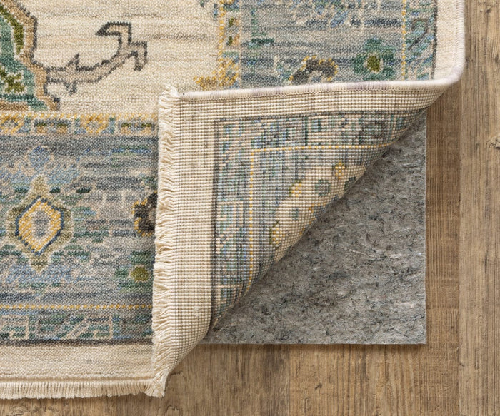 A corner of a patterned rug with blue, beige, and yellow designs and fringed edges is flipped over on a wooden floor, revealing the eco-friendly Oriental Weavers Luxehold / 0005E Rug Pad beneath it. The cushioned comfort of the rug pad ensures softness underfoot.