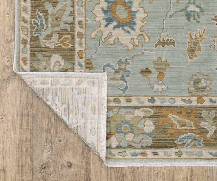 A corner of the Oriental Weavers Maeve / MAE06 Rug is folded over on a wooden floor, revealing the underside. This rug showcases Persian-inspired floral and geometric designs in blue, green, tan, and cream tones.
