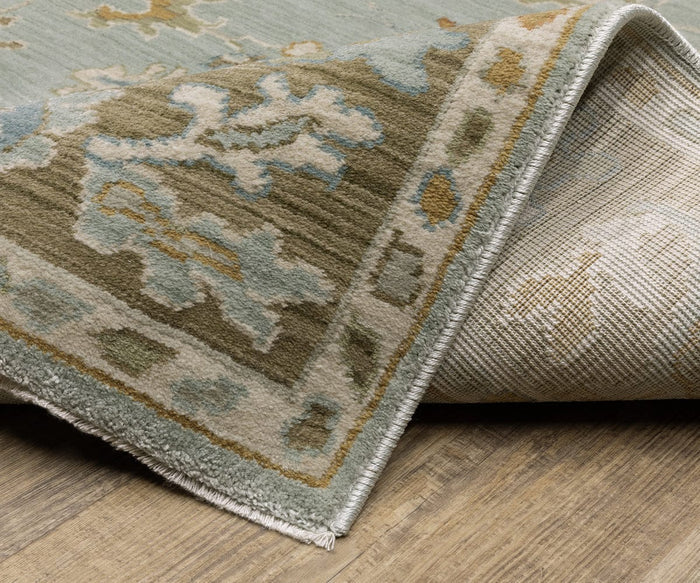 A corner of the Oriental Weavers Maeve / MAE06 Rug is folded over on a wooden floor, showcasing its detailed design in muted green, beige, and blue tones. Persian-inspired and crafted from space dyed polypropylene, this rug combines durability with subtle color variation.
