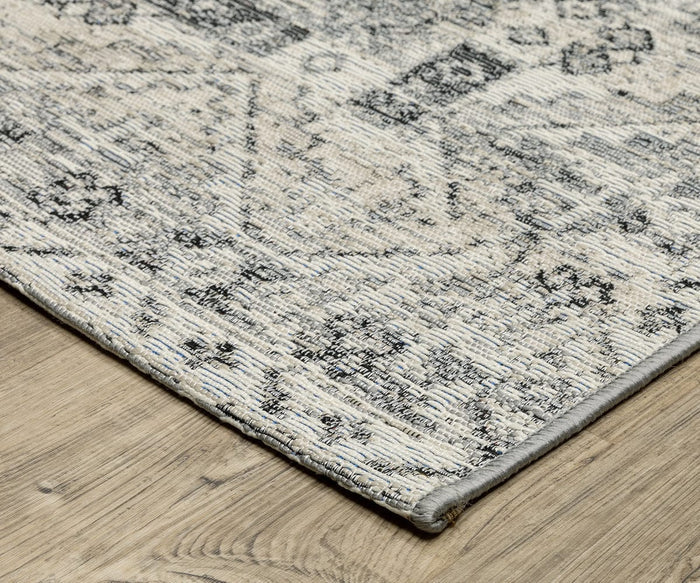 Close-up of a corner of the Oriental Weavers Naples / 1346Z Rug, featuring a distressed gray and beige pattern on a light wood floor. This polypropylene area rug has a textured, woven appearance and neatly finished edges, suitable for both indoor and outdoor use.
