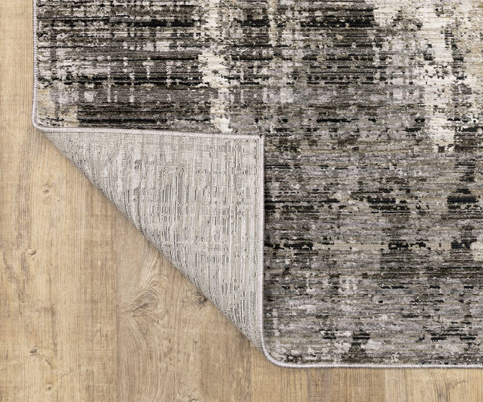 A corner of the Oriental Weavers Nebulous / 4151N Rug, featuring a sophisticated gray and black pattern, is folded over on a light wooden floor to showcase its textured underside. Expertly power-loomed from polyester, this modern abstract rug delivers both style and durability for contemporary interiors.