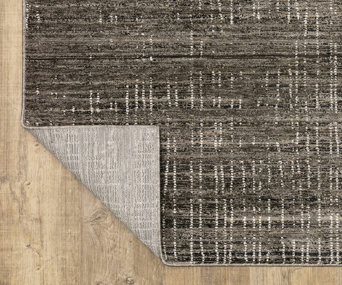 An Oriental Weavers Nebulous / 751D Rug, featuring a gray and black abstract pattern with white accents, is placed on a light wooden floor. One corner of this modern, space-dyed polyester area rug is flipped over to show its lighter non-slip backing and hint at its durable construction.