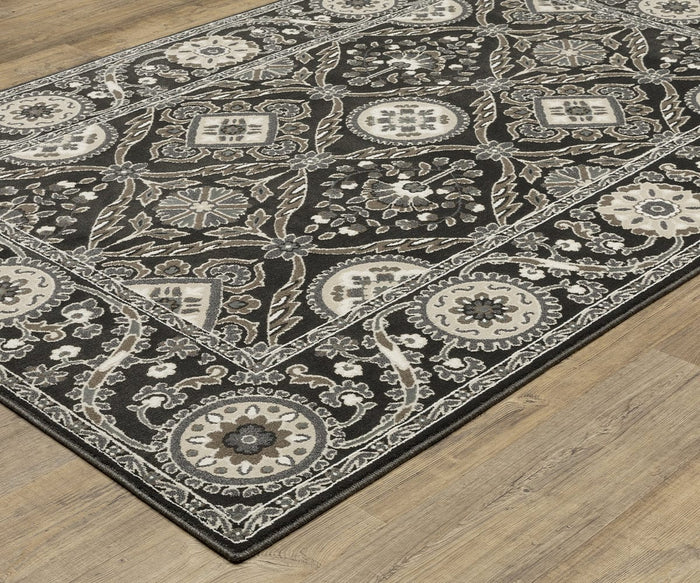 The Oriental Weavers Raylan / RAY05 Rug, featuring a rectangular shape, displays a black and gray floral and geometric pattern on a light wood floor. Its design showcases intricate borders and symmetrical motifs in beige, white, and gray tones with high/low construction for added texture.