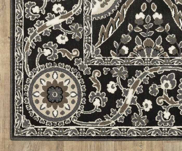 A close-up of the Oriental Weavers Raylan / RAY05 Rug, featuring intricate beige, gray, and white floral patterns with subtle geometric motifs on a decorative black background. The rug is displayed on a wooden floor, with a corner showcasing its detailed circular floral design.