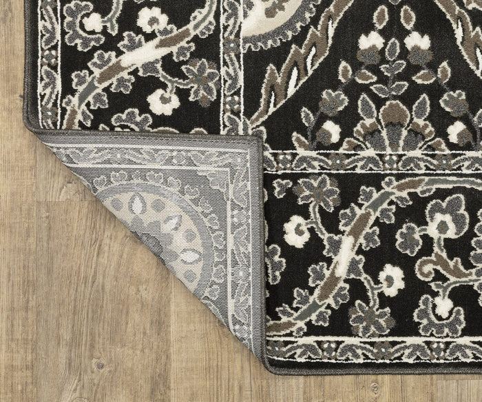 The Oriental Weavers Raylan / RAY05 Rug features a black and white floral pattern with subtle geometric motifs. One corner is folded over to reveal its gray underside, highlighting the rug’s textured high/low construction that adds depth and visual interest. Displayed on a light wood floor, this design creates a striking foundation for any space.