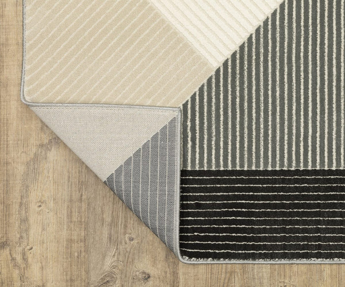 A close-up of a corner of the Oriental Weavers Raylan / RAY07 Rug on wooden flooring. The rug is folded to reveal beige, gray, and black sections with various stripe patterns and subtle geometric motifs, creating an eye-catching contrast in texture and design.