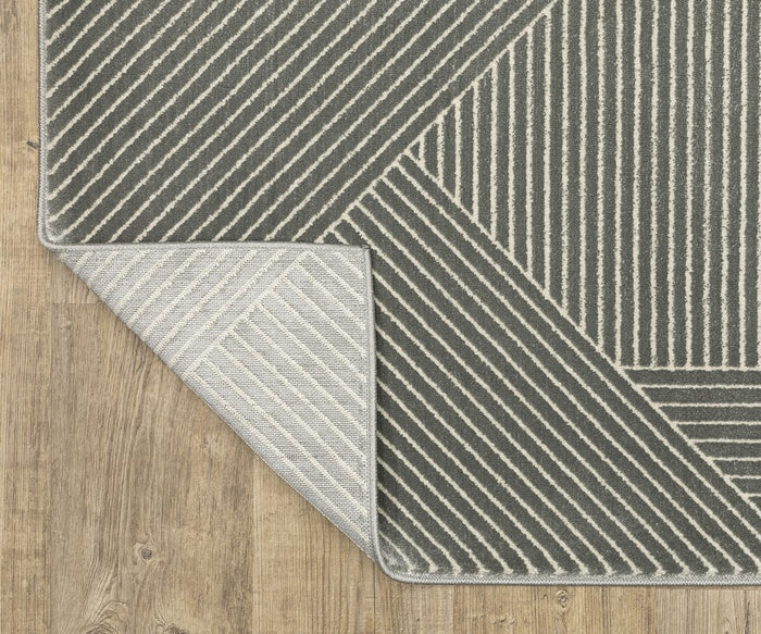 An Oriental Weavers Raylan / RAY08 Rug featuring geometric diagonal white and gray stripes is spread out on a wooden floor. One corner of this fade-resistant rug is flipped over to reveal the underside, which displays a lighter version of the same geometric pattern.