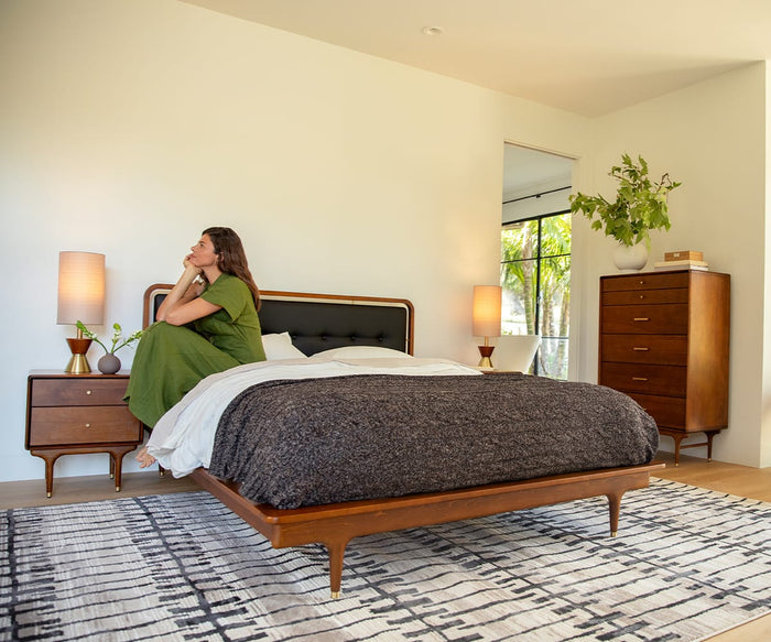 A woman in a green dress sits on a neatly made bed in a modern bedroom with wooden furniture, sunlight streaming through a window framed by greenery outside, and an Oriental Weavers Raylan / RAY11 Rug featuring a striking geometric motif that adds visual interest to the space.