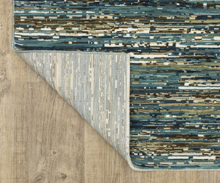 A corner of the Oriental Weavers Reed / RE01G Rug, crafted from space-dyed polypropylene, showcases abstract blue, green, and beige patterns as it folds over on a light wood floor, revealing the rug’s lighter underside with a similar design.