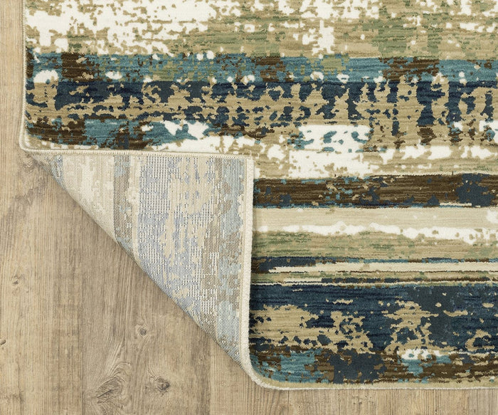 A corner of the Oriental Weavers Reed / RE02A Rug, featuring abstract blue, beige, green, and brown patterns, is folded over to reveal the underside of this textured space-dyed polypropylene rug resting on a light wood floor.