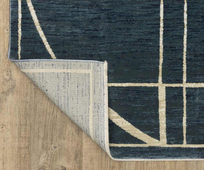 A corner of the Oriental Weavers Reed / RE04B Rug, in a dark blue and beige pattern, is flipped over to reveal the underside on a light wood floor. The textured rug features space-dyed polypropylene fibers for added depth and durability.
