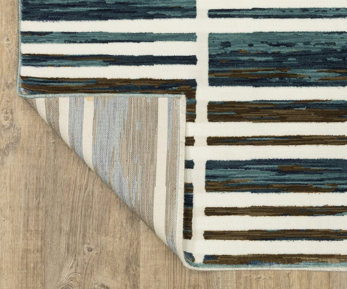 The Oriental Weavers Reed / RE05A Rug showcases blue, brown, and cream horizontal stripes in a modern design. Crafted from space-dyed polypropylene for enhanced depth and durability, this textured rug adds contemporary style and subtle dimension to any room. Displayed on a wooden floor with one corner folded back to reveal the muted linear pattern of the underside, it combines visual interest with everyday practicality.